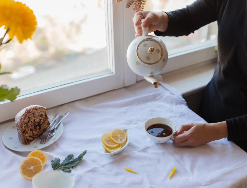 Person pouring tea by window with cake